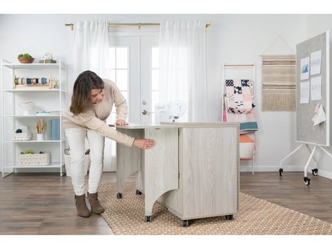 A woman opens a light wood sewing cabinet with a sewing machine on top.