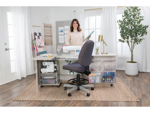 A woman stands smiling behind a sewing cabinet with a sewing machine and supplies.
