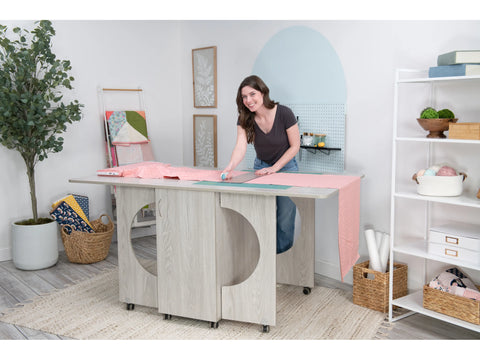 A woman cuts fabric on a light wood Koala Cutting Table with storage and wheels.