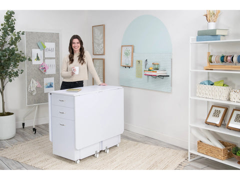 A woman stands next to a white Koala Cutting Table with two drawers and drop leaves.