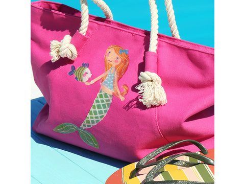 Pink canvas tote bag with a mermaid and fish design, next to striped flip-flops.