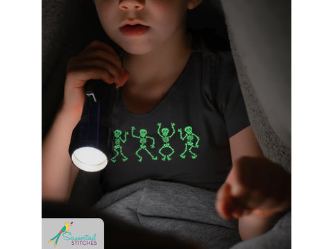 A child holds a flashlight, illuminating a black shirt with four glowing green dancing skeletons.