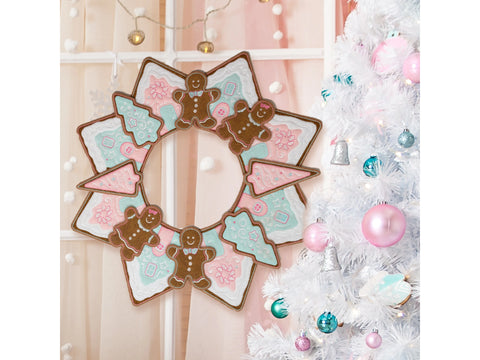 A freestanding gingerbread wreath hangs on a window next to a white Christmas tree.