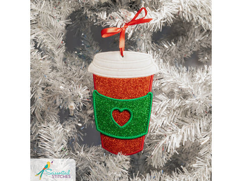 A glittery red coffee cup ornament with a green heart-shaped sleeve hangs on a white Christmas tree.