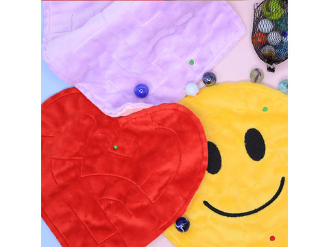 Three plush maze game boards, one purple, one red heart-shaped, and one yellow smiley face, are displayed with marbles and a mesh bag of marbles.