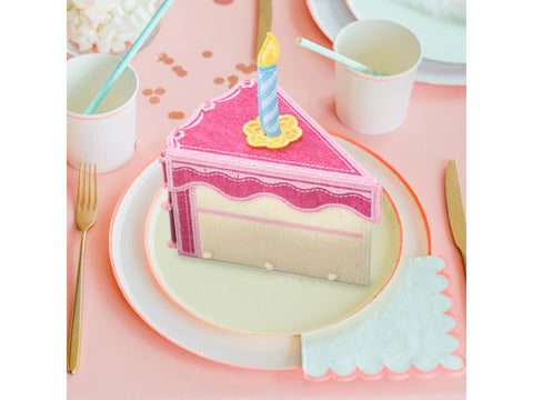A pink fabric cake box with a candle on top sits on a plate with party supplies.