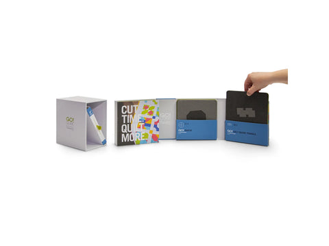 A hand places a dark gray die into a blue box, next to a book and other dies.