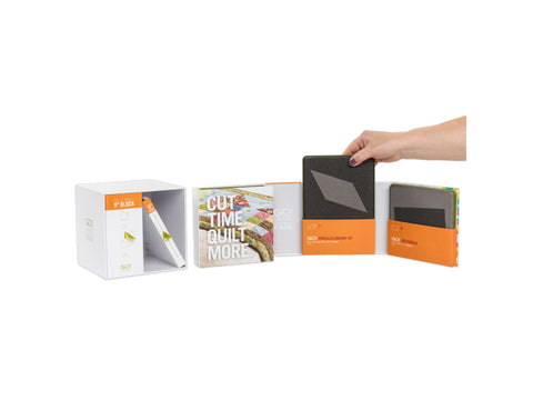 A hand places a dark gray parallelogram die into an orange box, next to a white box with a "Cut Time Quilt More" book.