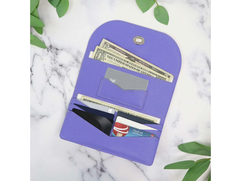 A purple faux leather wallet with cash and cards inside, on a marble surface with green leaves.