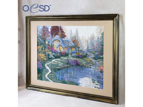 A framed embroidery of a cottage with glowing windows, a winding path, and a pond.