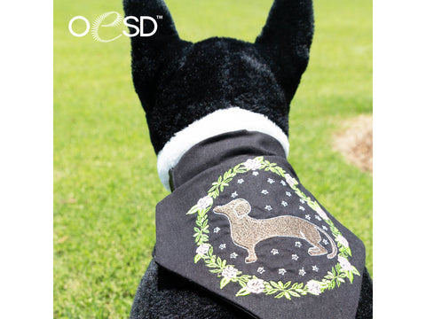 A black dog wears a bandana with an embroidered dachshund in a floral wreath.