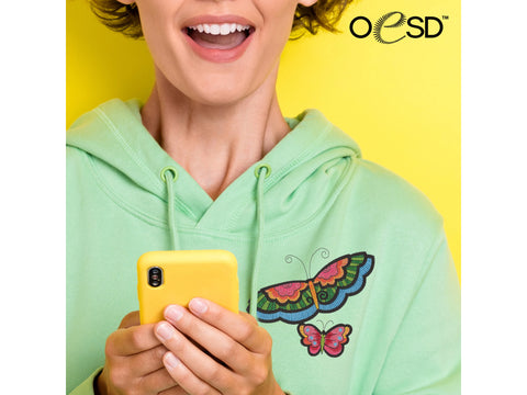 A person in a light green hoodie holds a yellow phone, showcasing two colorful butterfly embroideries.