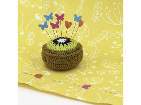 A kiwi-shaped pincushion holds colorful butterfly and heart-shaped flat head pins on a yellow fabric.