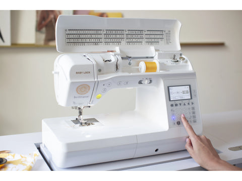 A white Baby Lock Brilliant sewing machine with a yellow spool of thread and a hand pressing a button.