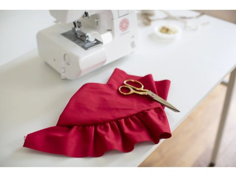 A Baby Lock Vibrant serger sits on a white table next to red ruffled fabric and gold scissors.