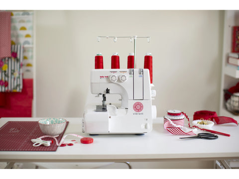 A white Baby Lock Vibrant serger with red thread spools sits on a white table with sewing supplies.