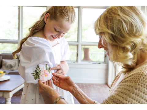 A young girl smiles as an older woman adjusts the embroidered sleeve of her dress.