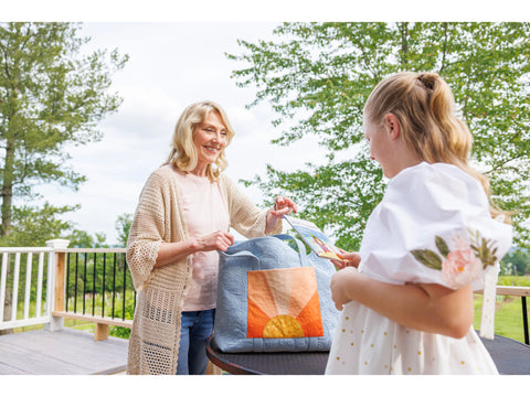 A woman and a girl on a deck with a light blue quilted bag featuring a sun design.