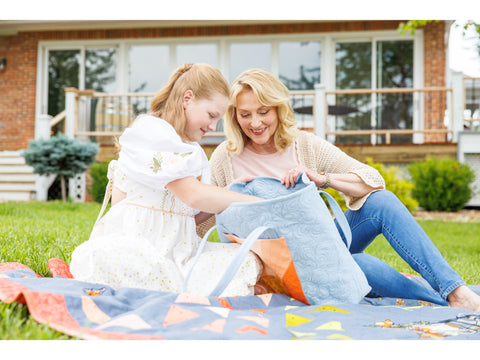 Two women on a quilt on grass, one holding a light blue quilted bag.