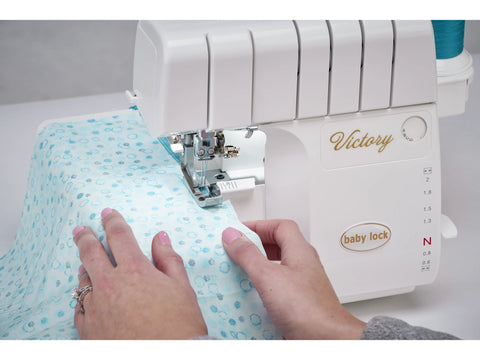 A white Baby Lock Victory serger with a light blue patterned fabric being sewn.