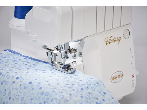 A white Baby Lock Victory serger with blue thread and patterned fabric.