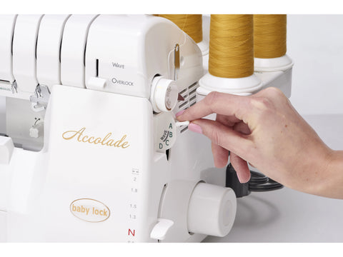 A hand adjusts settings on a white Baby Lock Accolade serger with gold thread spools.