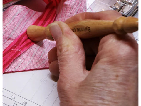 A hand holds a wooden stiletto and pressing tool against pink striped fabric.
