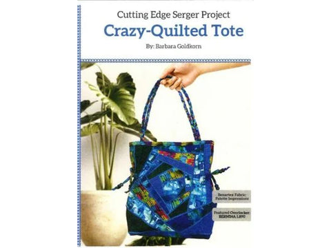 A hand holds a blue crazy-quilted tote bag with colorful accents, next to a potted plant.