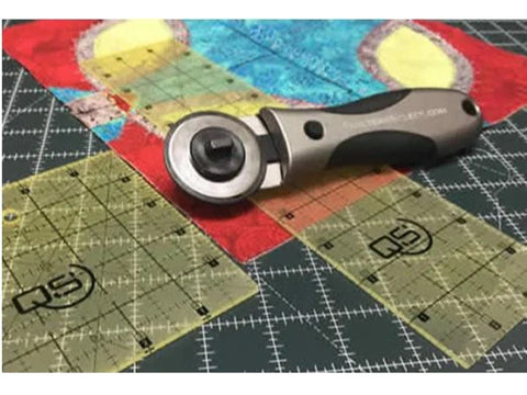 A Quilters Select 28mm Deluxe Rotary Cutter rests on a cutting mat with fabric and rulers.