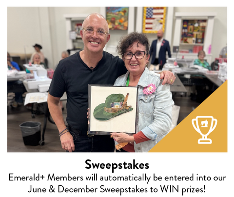 Two people smile, one holding a framed artwork of a green hat with feathers.