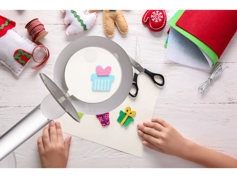 A child's hands are visible on a white wooden table, crafting felt Christmas decorations with a Halo Go Rechargeable Magnifier.