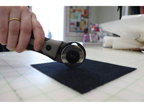 A hand holds a gray and black rotary cutter over a piece of dark fabric on a cutting mat.