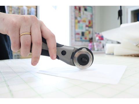 A hand holds a Quilters Select 45mm Deluxe Rotary Cutter over white fabric on a cutting mat.