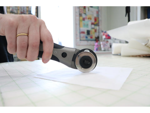 A hand holds a gray and black rotary cutter over white fabric on a grid mat.