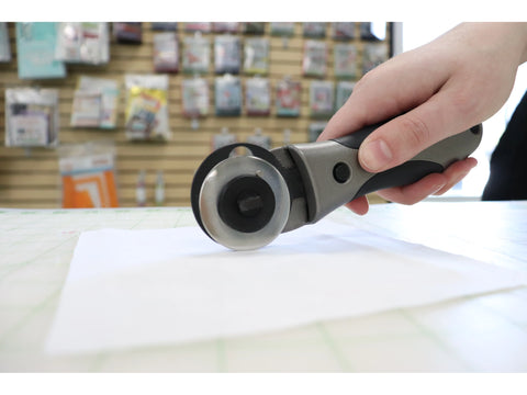 A hand holds a gray and black rotary cutter over a piece of white fabric.