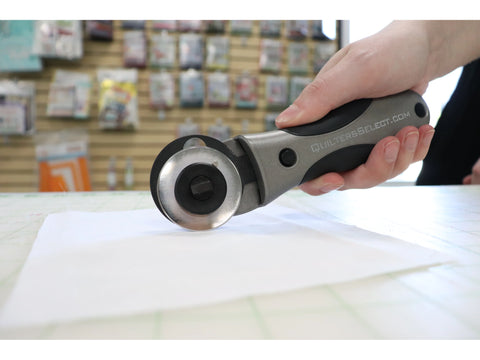A hand holds a gray and black rotary cutter over white fabric on a cutting mat.