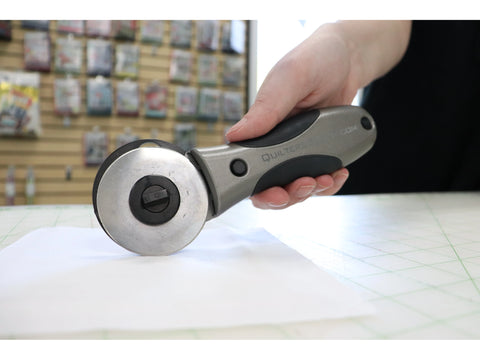 A hand holds a gray and black Quilters Select 60mm Deluxe Rotary Cutter on white fabric.