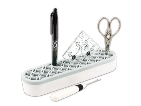 A gray and white organizer holds a black pen, a small clear ruler, and silver scissors, with a seam ripper in front.