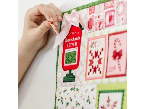 A hand places a small orange tag into a red "Dear Santa Letters" mailbox on a Christmas-themed quilt.