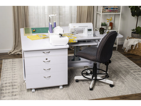 A white sewing table with two sewing machines, a rolling storage chest, and an office chair.