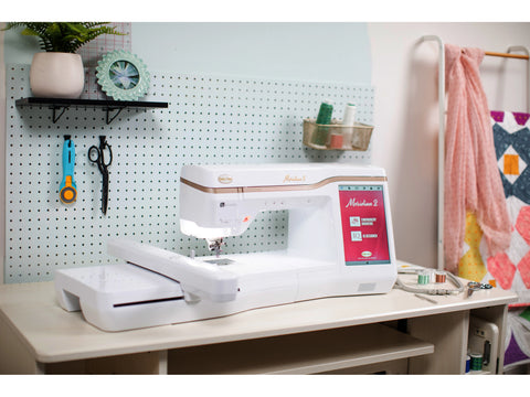 A white Baby Lock Meridian 2 embroidery machine sits on a light wood desk in a craft room.