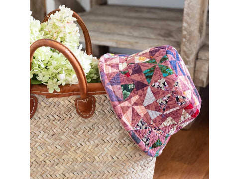 A woven straw bag with leather handles holds white hydrangeas and a colorful patchwork bucket hat.