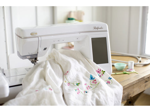 A white Baby Lock Pathfinder embroidery machine is actively stitching a floral design on white fabric.