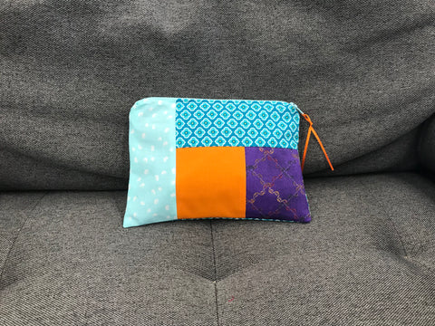 A colorful patchwork pouch with a zipper and orange pull tab rests on a gray textured surface.
