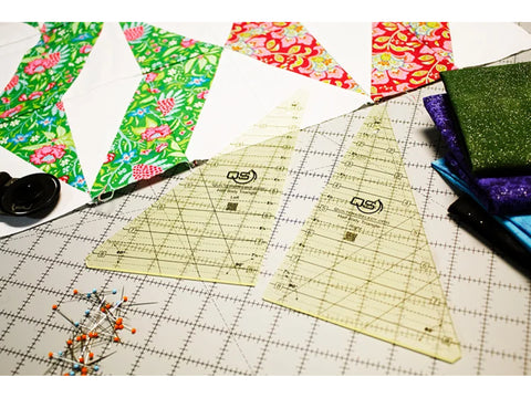 A set of two clear, triangular Quilters Select rulers rests on a white cutting mat with quilt blocks.