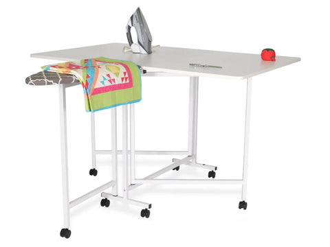 A white cutting and ironing table with a colorful quilt, iron, scissors, and pin cushion.