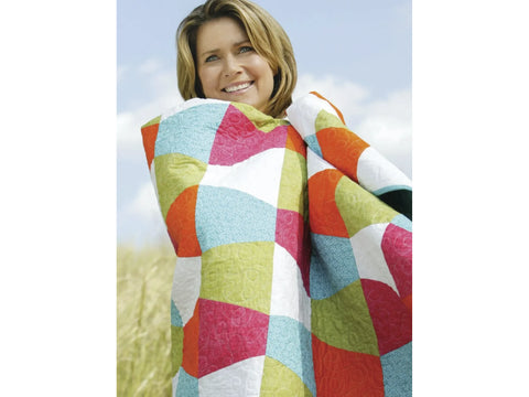 A smiling woman wrapped in a colorful patchwork quilt with a blue sky background.