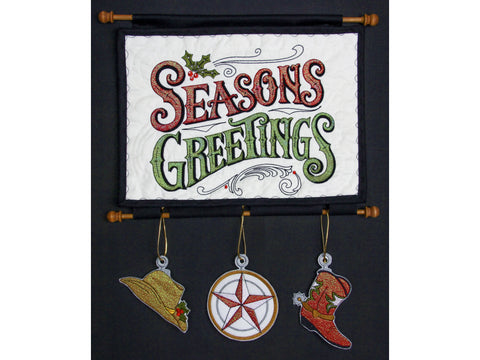A black wall hanging with a white embroidered "Seasons Greetings" sign and three western-themed ornaments.