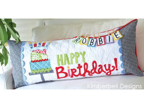 A white bench pillow with "Happy Birthday Robbie!" embroidered on it, featuring a cake and candles.