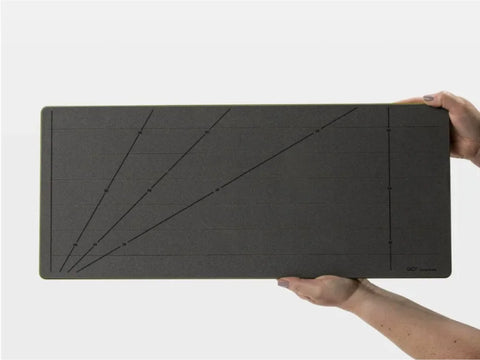 Two hands hold a dark gray cutting mat with measurement lines and angles.